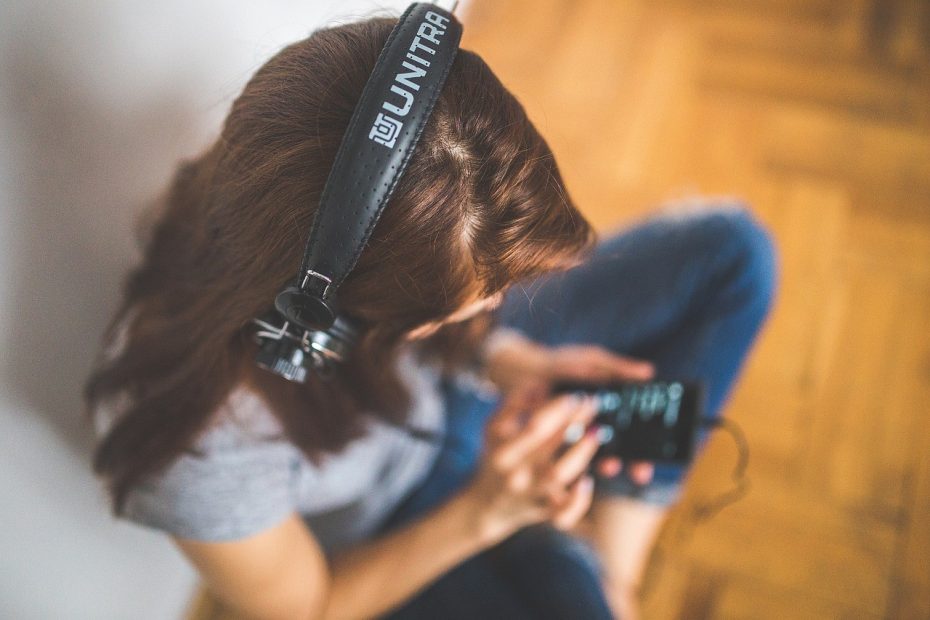 a woman sitting on the wooden floor, listening to a podcast with her headphones connected to her phone